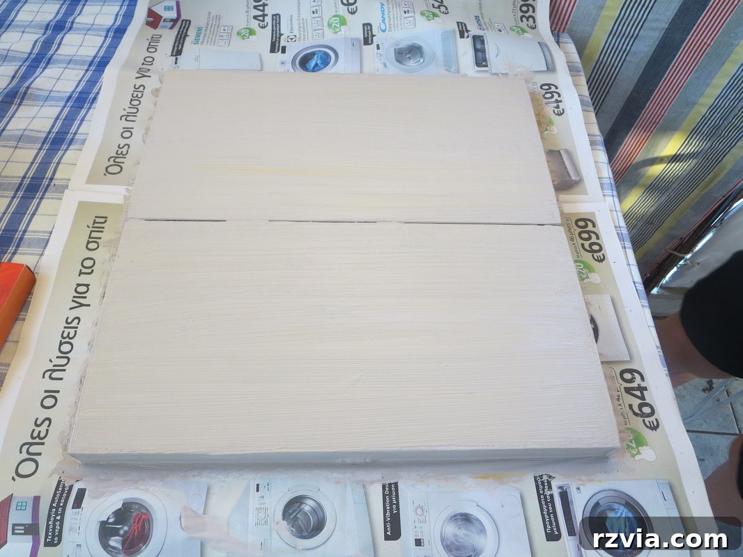 A close-up shot of hands applying white chalk paint to the old laminate box. The chess pattern is still faintly visible beneath the first coat, illustrating the painting process. The image focuses on the texture and coverage of the chalk paint as it transforms the surface.