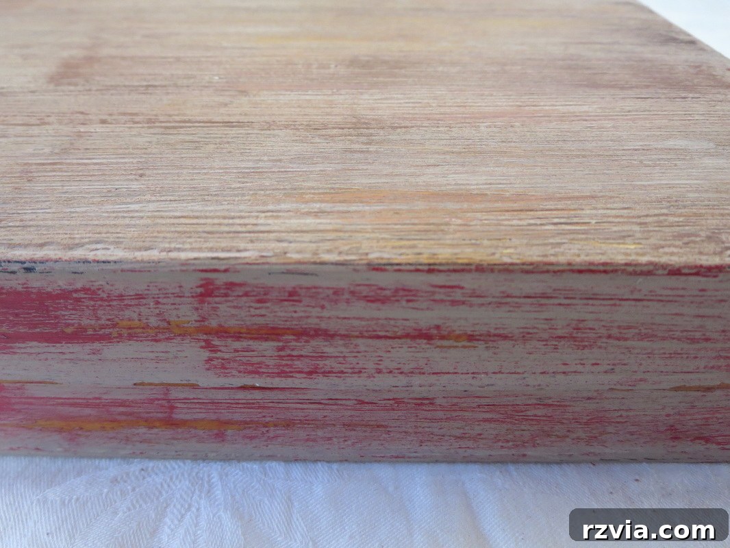 A child's hand (the 'little helper') applying distressed red paint to the side of the textured laminate box. The red paint is being lightly brushed, allowing the underlying wood-like texture to show through, creating an aged and vibrant accent.