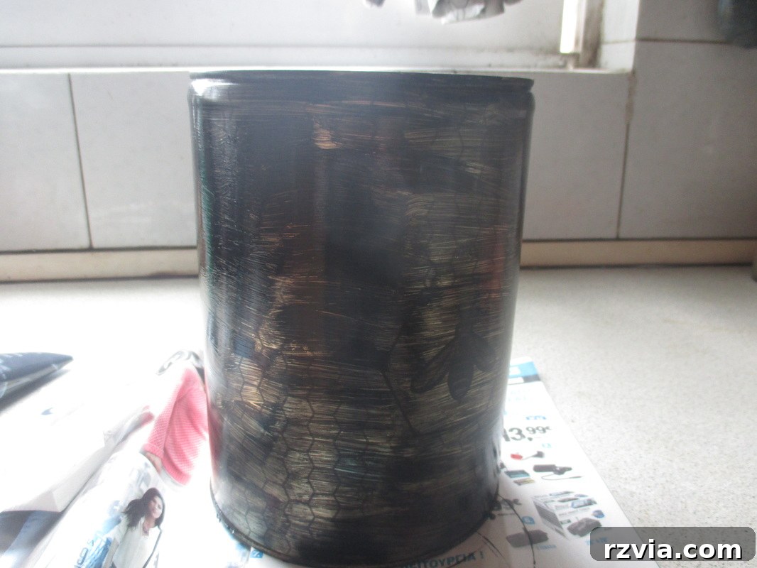 The honey tin painted black, a crucial step in creating a distressed, rustic kitchen utensil holder. This solid base prepares the tin for further detailing and adds a modern touch to the DIY project.