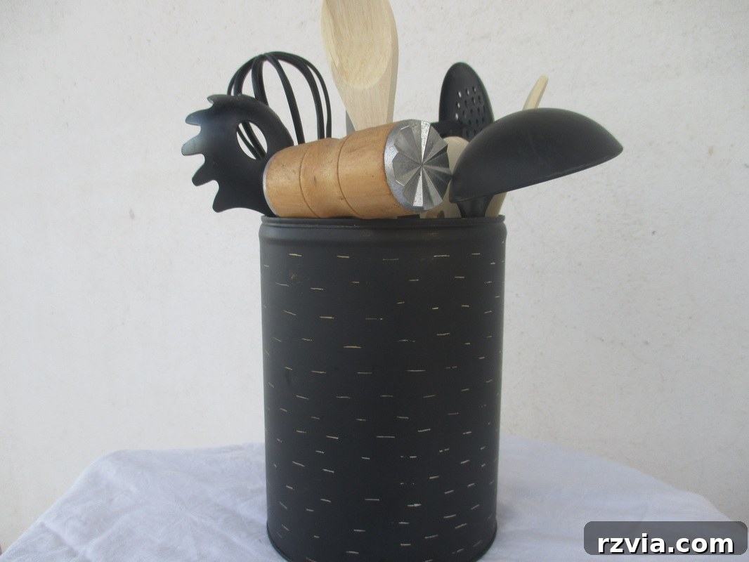 Another view of the DIY imperfect kitchen utensil holder, an upcycled honey tin, featuring a distressed black finish and holding a variety of kitchen tools. Ideal for farmhouse or rustic kitchen decor.