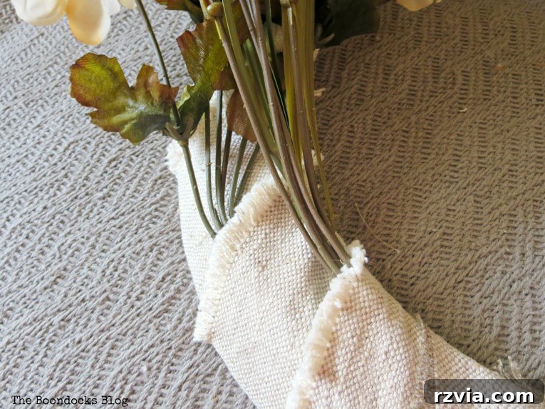 A hand gently placing faux fall flowers and wheat grass into the fabric loops of the wrapped wire wreath form, adding vibrant color and texture.