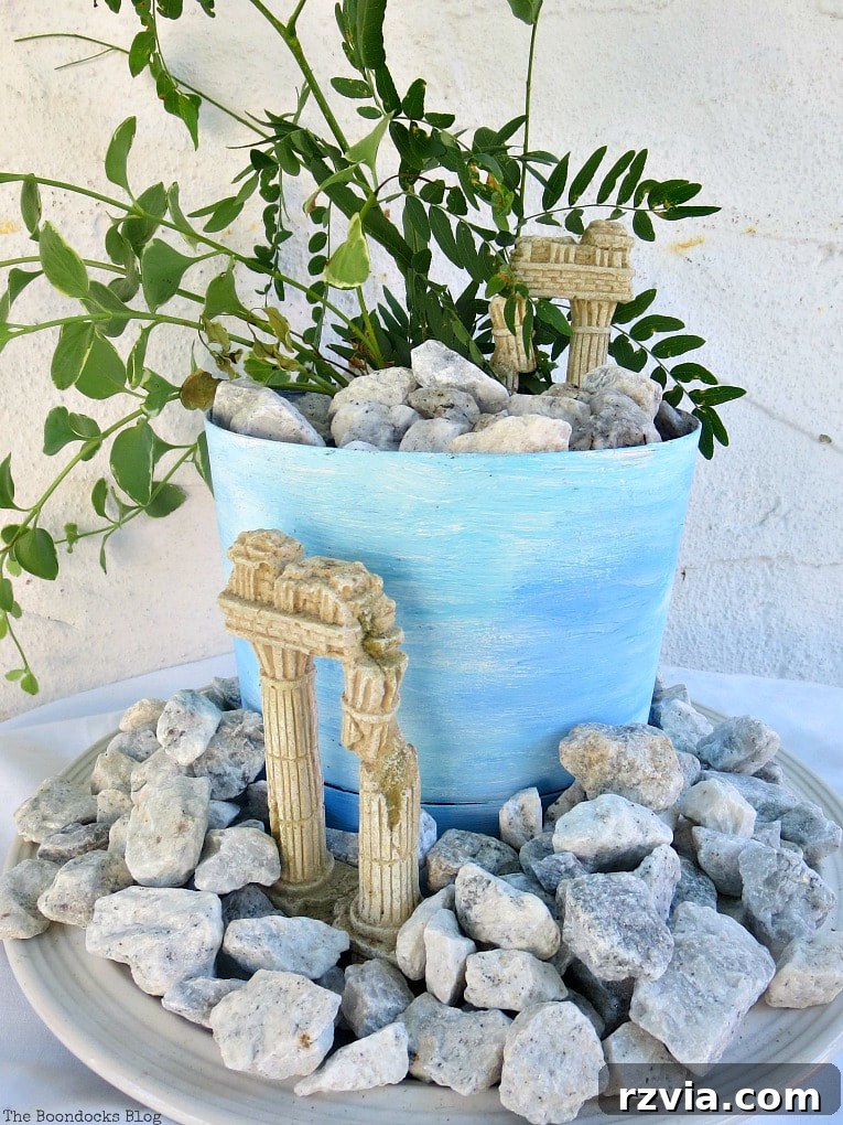 Seaside Style from Salvaged Treasures 16 A complete beachy planter vignette featuring a blue and white painted planter, vinca vine, and ancient 'ruins' amidst pebbles, exuding Mediterranean charm.