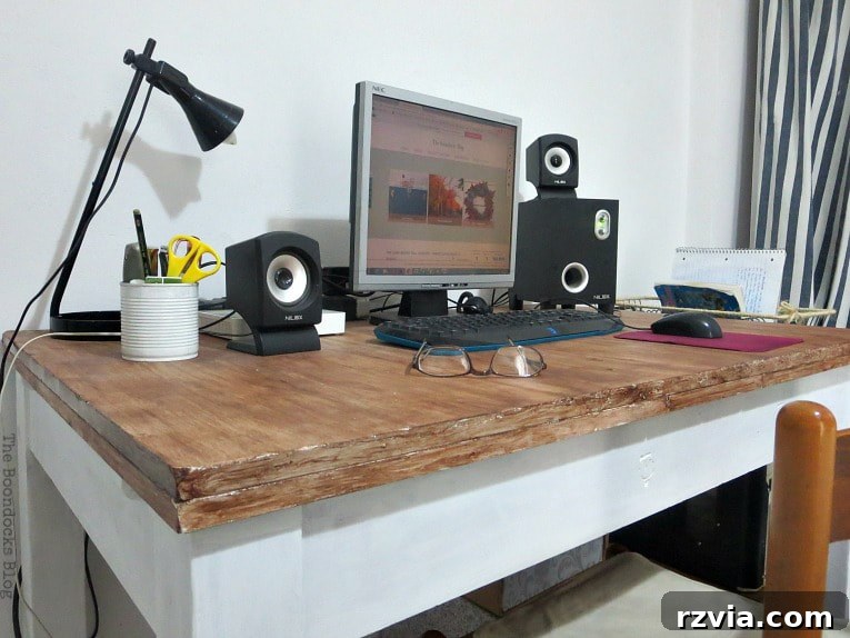A tastefully arranged wooden desk serving as a home office, featuring a computer, a sleek black lamp, glasses, speakers, and a minimalist pen holder, all contributing to a functional yet stylish workspace.