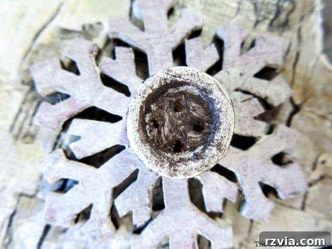 A hand holding a newly painted snowflake embellishment with a brown distressed button, ready to be attached to the star.