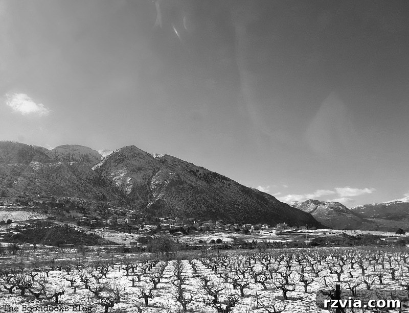 The White Crowns of Greece 6 Black and white photograph of vineyards (raisin fields) near a small, snow-dusted Greek mountain village, depicting rural life at high altitude.
