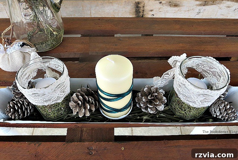 The completed first version of the Christmas centerpiece featuring a white chicken feeder, olive leaves, rosemary, pinecones, and a green ribbon around the candle. Shows the natural and rustic aesthetic. Photo by www.theboondocksblog.com