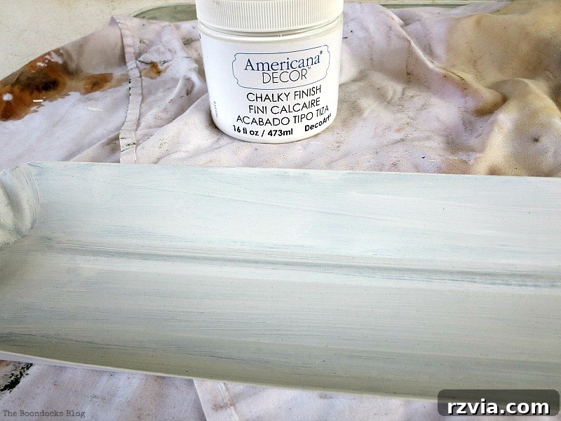 Hands painting a chicken feeder white with American Decor chalky finish paint, preparing it for a DIY Christmas centerpiece. Shows the transformation process. Photo by www.theboondocksblog.com