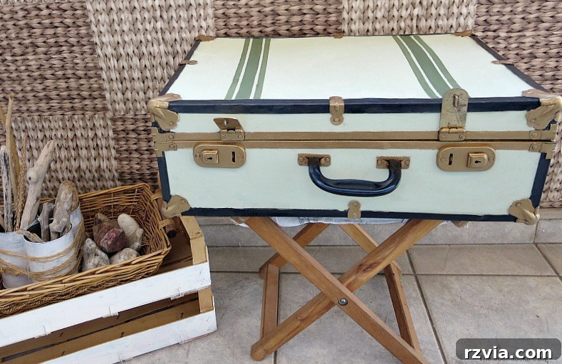 Breathing New Life into Old Luggage 16 The finished vintage suitcase, adorned in white, gold, soft olive green, and black, elegantly positioned on a brown table next to a rustic basket filled with driftwood and rocks, showcasing its decorative potential.