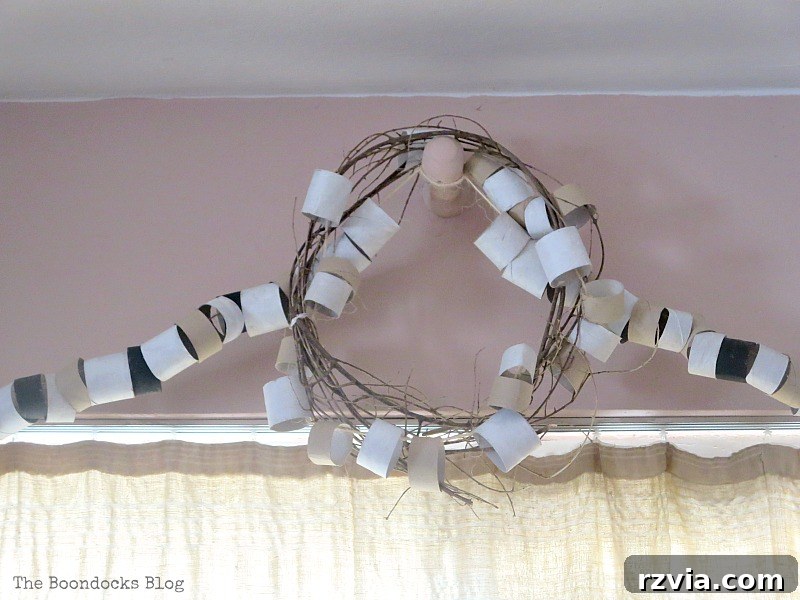 A rustic and charming wreath, skillfully crafted from repurposed paper rolls, intertwined with natural twigs and twine. This decorative piece was specifically designed to cover a central, unsightly bracket, adding a focal point to the decor.
