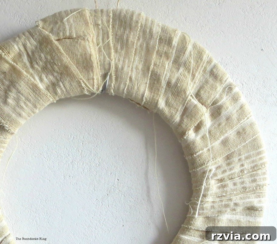 A hand wrapping a cardboard wreath base with strips of fabric, illustrating the initial steps of a DIY spring wreath project.