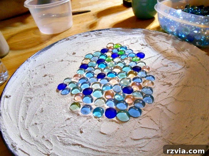 Craft Your Own Sea Glass Mosaic Table 6 Hands carefully arranging various flat-sided glass marbles and sea glass onto the freshly applied grout on the pizza pan, beginning the mosaic design.
